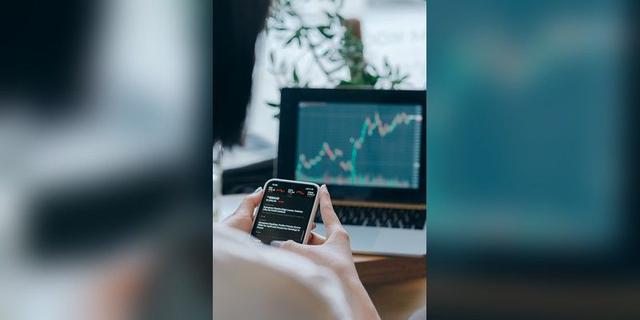 Person using a smartphone beside a laptop displaying a market chart