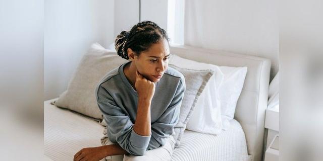 Young woman sitting thoughtfully in a bedroom, used as neutral illustrative lifestyle imagery.