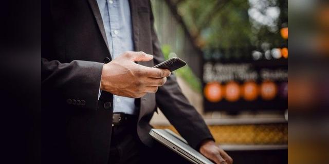 Professional using a smartphone while holding a laptop outdoors