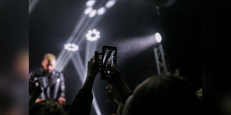 Concert crowd and silhouetted performer under stage lights with a person filming on a smartphone, used as illustrative music imagery