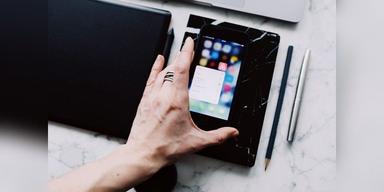 A hand using a smartphone beside a desk, tablet and writing tools, shown as illustrative mobile-device imagery.