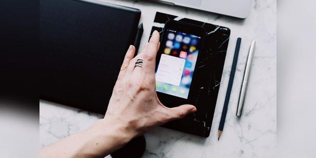 A hand using a smartphone beside a desk, tablet and writing tools, shown as illustrative mobile-device imagery.
