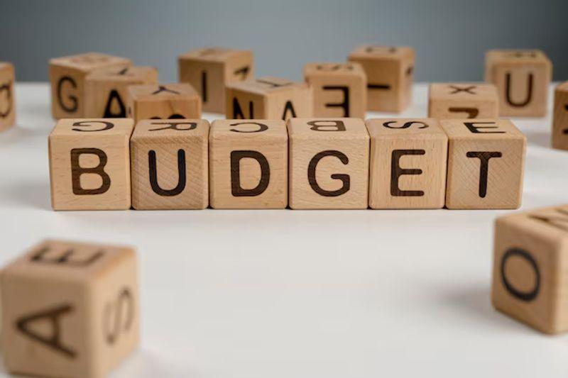 Person reviewing a monthly budget on a laptop with a calculator, notebook and household bills on a desk.