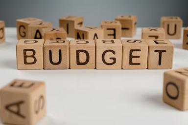 Person reviewing a monthly budget on a laptop with a calculator, notebook and household bills on a desk.