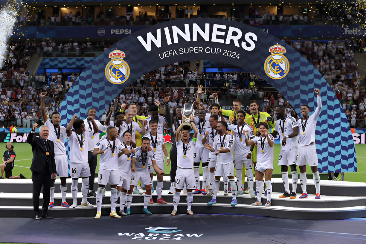 Real Madrid players celebrating on the podium after winning the UEFA Super Cup 2024, holding the trophy with confetti falling around them.