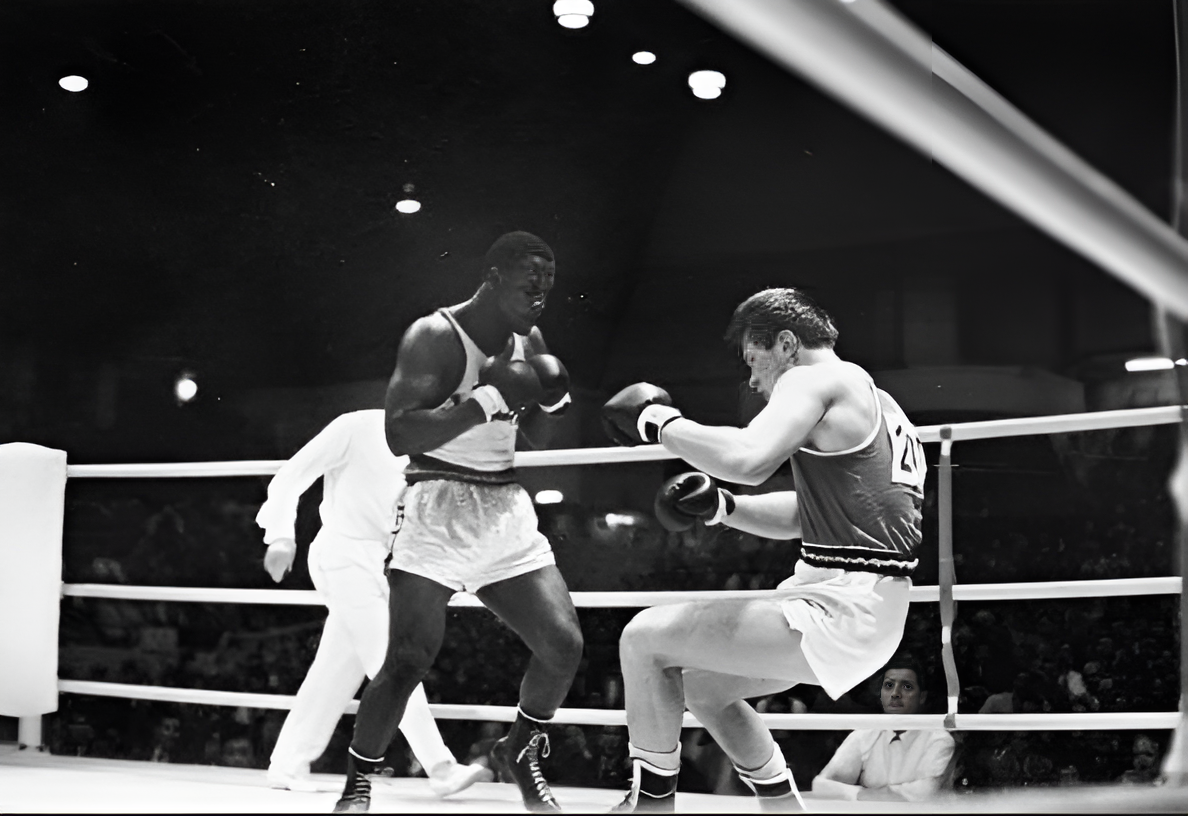 Black and white photo of Nigerian boxer Nojim Maiyegun in the ring during a match at the 1964 Tokyo Olympics, where he won a bronze medal. Maiyegun is seen standing in a defensive posture, while his opponent crouches defensively with raised gloves.
