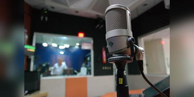 Studio microphone in a recording room with a blurred person behind the glass, used as illustrative radio-broadcast imagery.