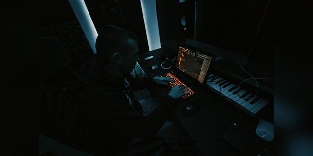 Music producer working at a laptop and keyboard in a dark studio, used as illustrative music-production imagery.