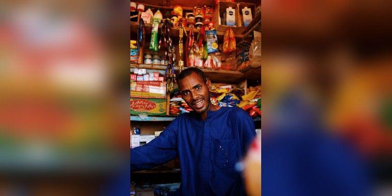 A Nigerian shopkeeper stands in a grocery store with shelves of packaged goods behind him.