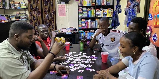Six people play a tabletop word game at a table surrounded by board games in Nigeria.