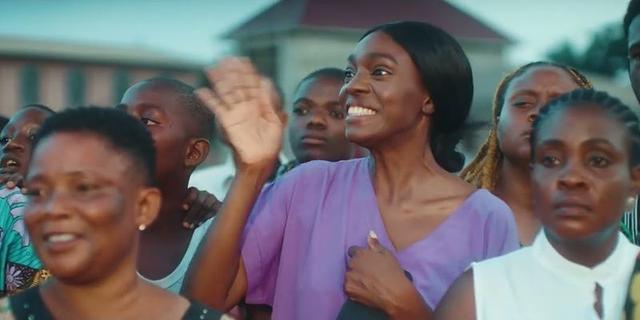 A woman in purple waves while standing among people in a scene from Ikenga: The Last Festival.