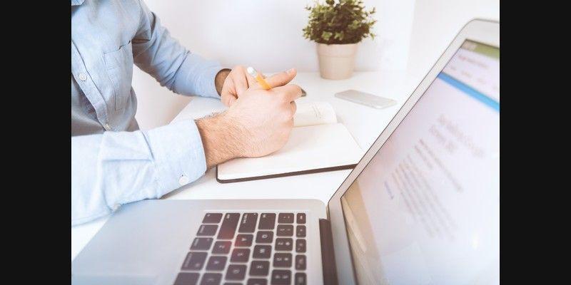 A businessman sits at a desk working on an open laptop while writing notes in a notebook.